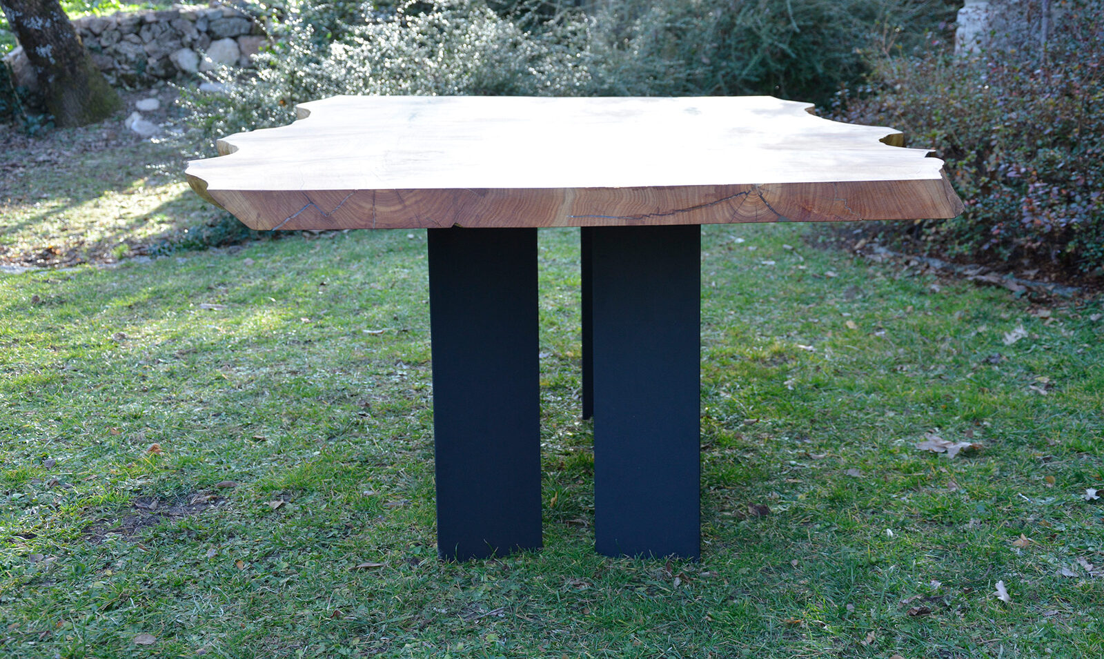 Mesa de tablero irregular con dos apoyos oscuros, en un jardín